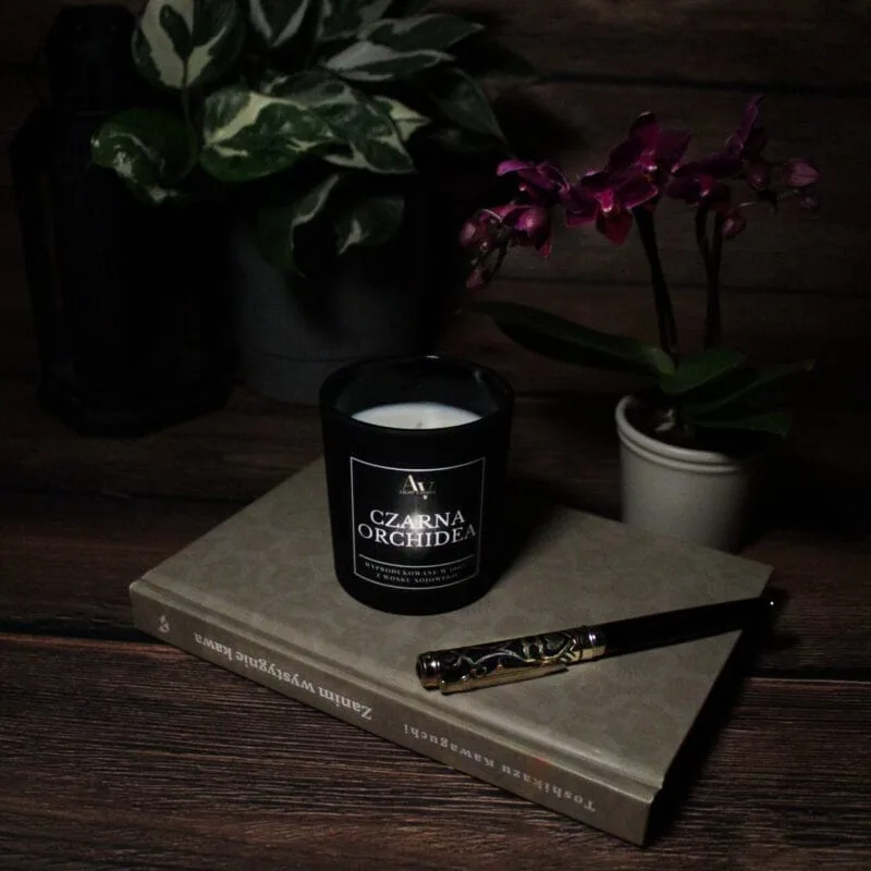 Świeca AV Light Candle w zapachu Czarna Orchidea, w matowym szkle, w otoczeniu eleganckich, ciemnych kwiatów, tworząca luksusowy klimat.