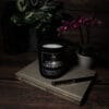 Świeca AV Light Candle w zapachu Czarna Orchidea, w matowym szkle, w otoczeniu eleganckich, ciemnych kwiatów, tworząca luksusowy klimat.