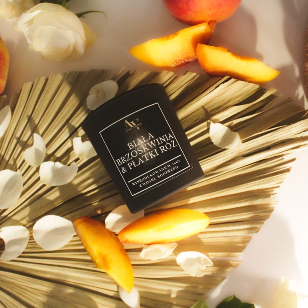 Świeca sojowa AV Light Candle Biała Brzoskwinia & Płatki Róż 130 ml – kolekcja Ember, ułożona na wachlarzu z liści palmowych, w otoczeniu brzoskwiń i białych płatków róż