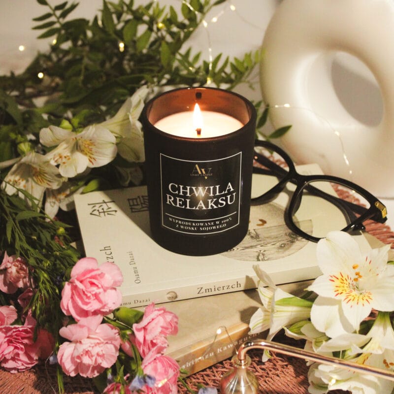 Zapalona świeca Chwila Relaksu Sense 200 ml na książkach wśród białych lilii i różowych goździków.