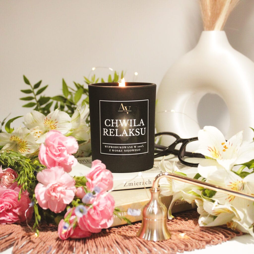 Świeca zapachowa Chwila Relaksu Sense 200 ml w czarnym szkle, otoczona kwiatami i książkami.
