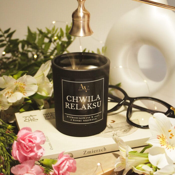 Świeca zapachowa Chwila Relaksu Sense 200 ml zapalona, otoczona kwiatami i okularami, na tle białej ceramicznej dekoracji.