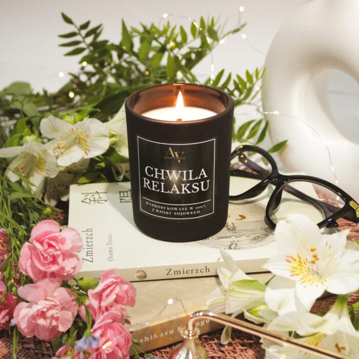 Zgaszona świeca Chwila Relaksu Sense 200 ml z unoszącym się dymem, w eleganckim otoczeniu kwiatów i książek.