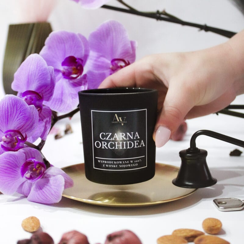 Zbliżenie świecy Czarna Orchidea AV Light Candle trzymanej w dłoni, z tłem z kwiatów orchidei.