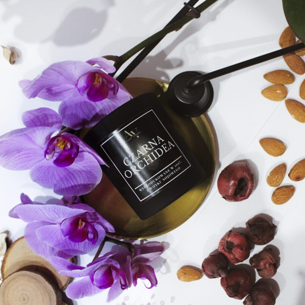 Czarna świeca sojowa AV Light Candle z kolekcji Ember o zapachu Czarnej Orchidei, ozdobiona fioletową orchideą i złotą podstawką.