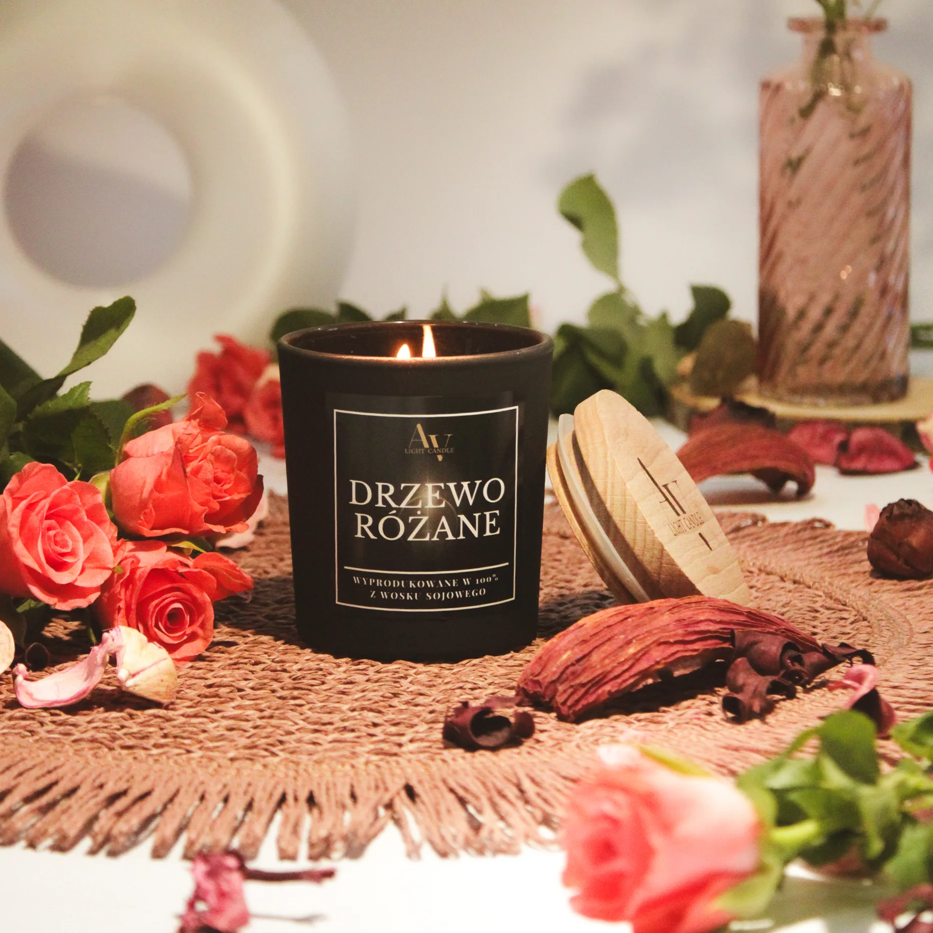 Zapalona świeca Drzewo Różane AV Light Candle z pokrywką, ustawiona na plecionym podkładzie