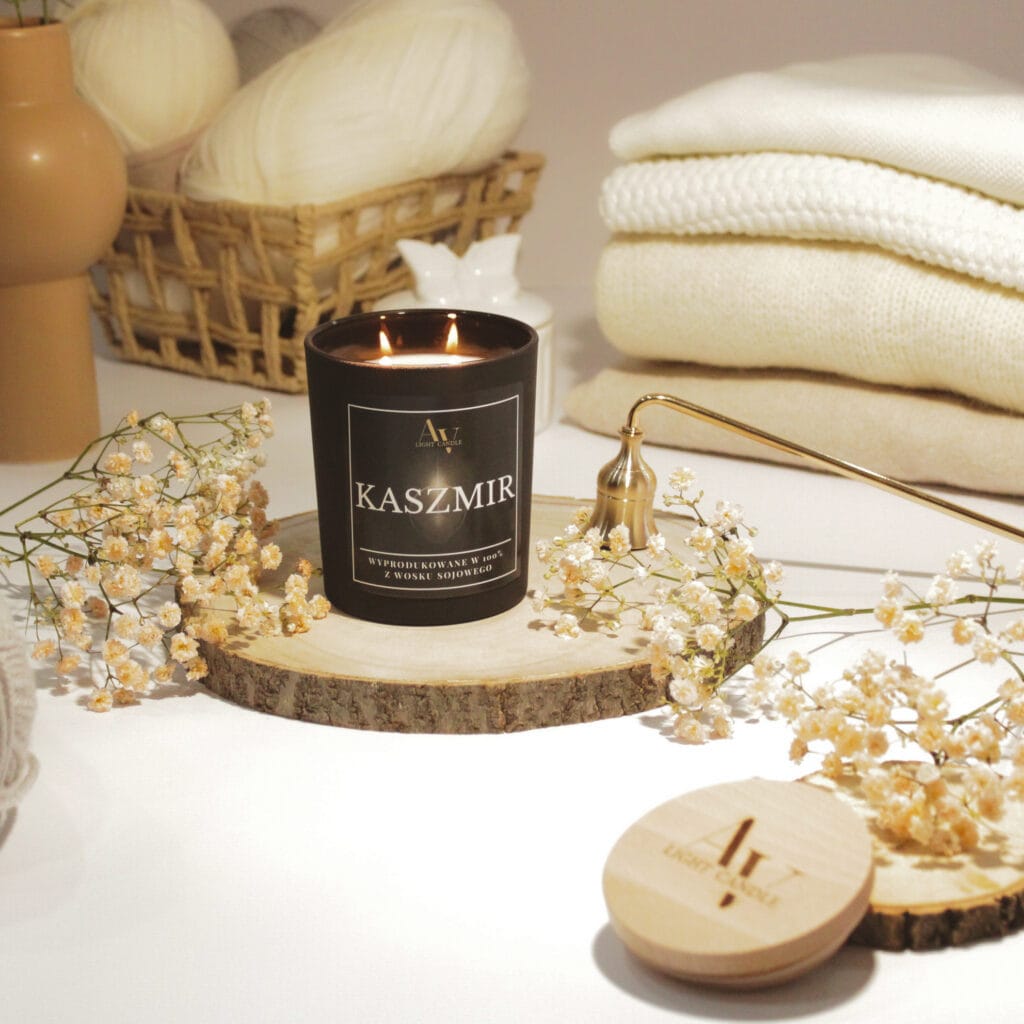 Świeca sojowa Kaszmir AV Light Candle 280 ml, paląca się na tle kremowych tkanin i kwiatów, elegancka aranżacja