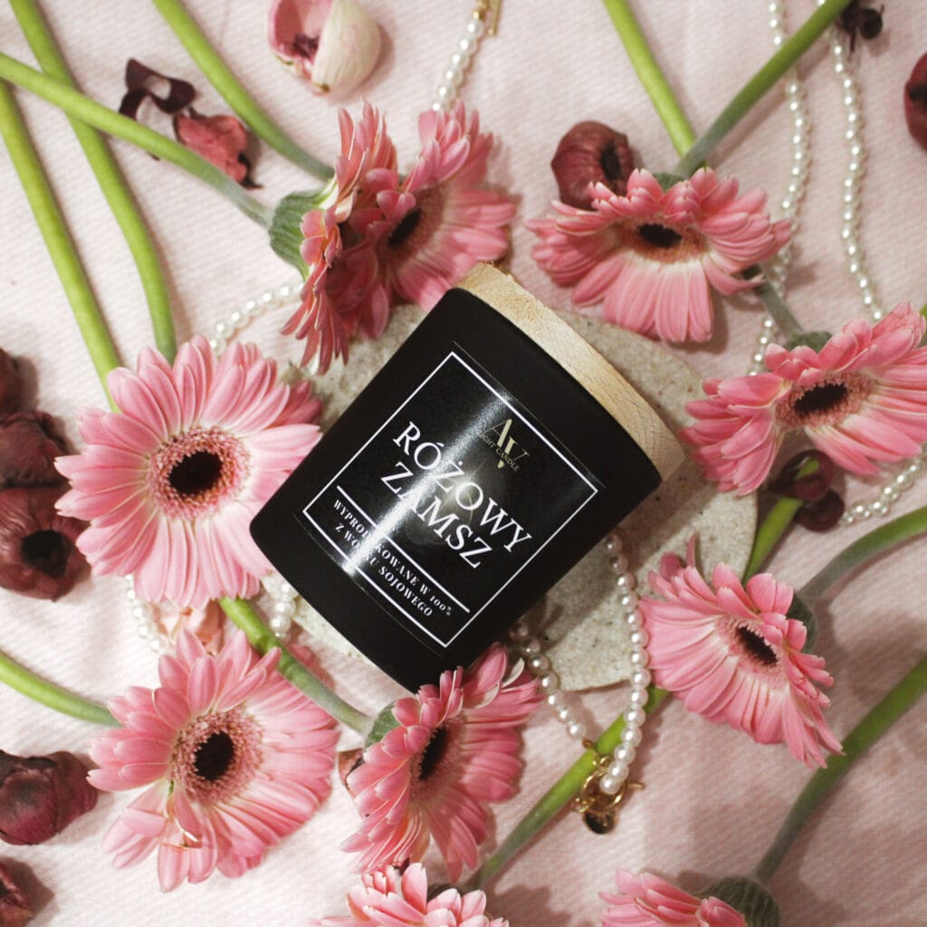 Świeca Różowy Zamsz Ember otoczona różowymi gerberami i perłami na jasnym tle