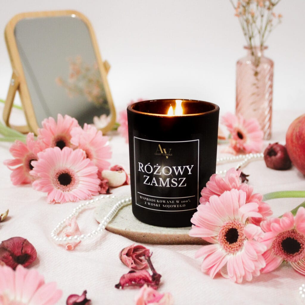 Czarna świeca sojowa AV Light Candle Różowy Zamsz 200 ml zapalona, w otoczeniu różowych kwiatów i pereł.