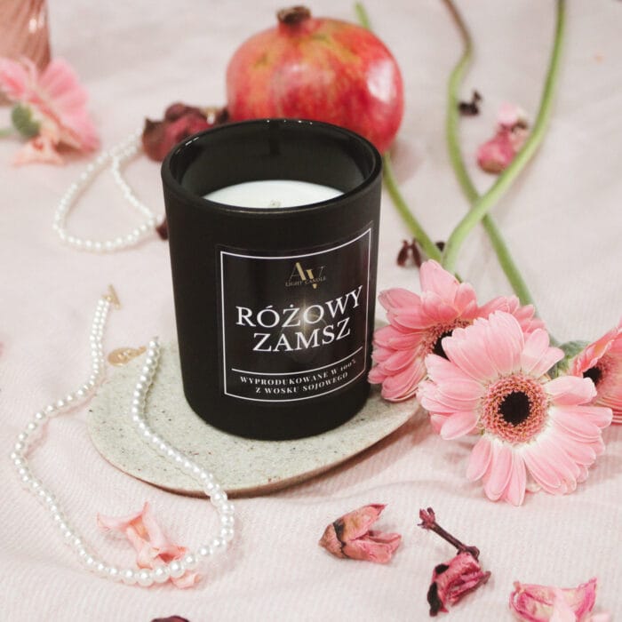 Świeca Różowy Zamsz 200 ml AV Light Candle otoczona różowymi gerberami i naszyjnikiem z pereł.